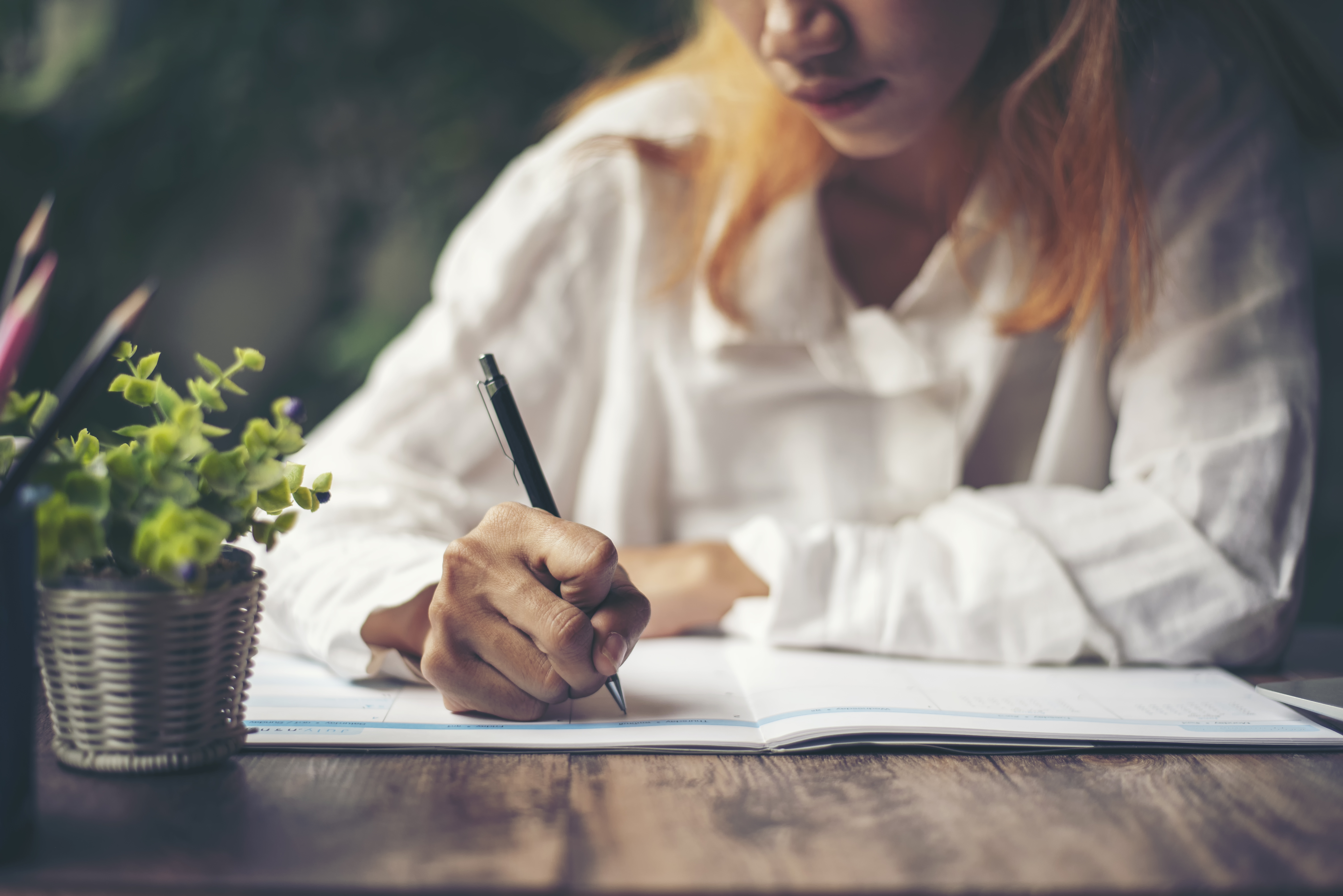 Image of a woman with peach hair and a white shirt writing in her notebook at a table