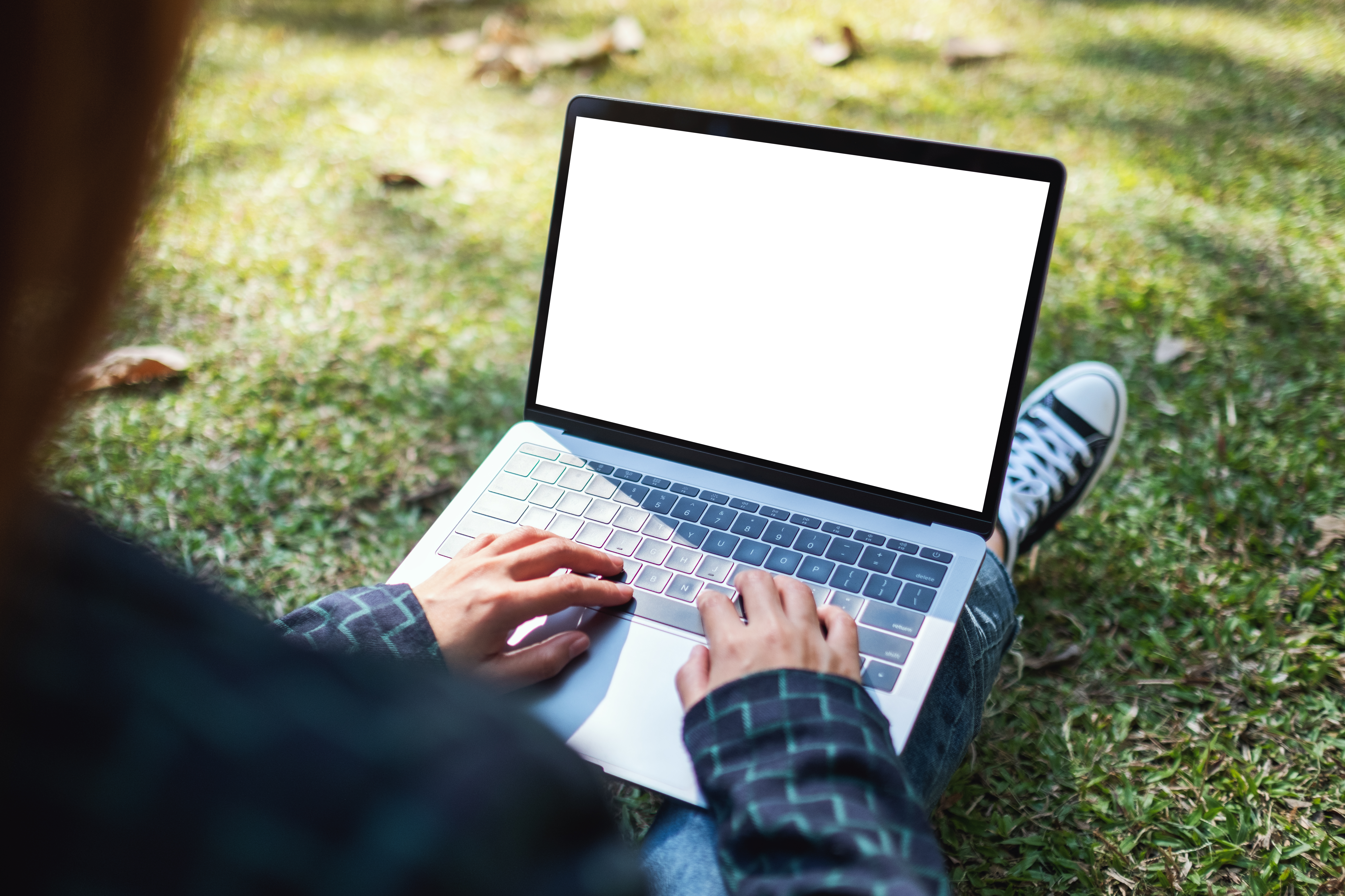 Image taken from behind a woman's shoulder as she sits on the grass, writing on her laptop